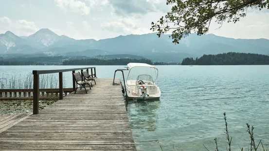 Holzsteg mit Bänken und weißem Boot auf klarem Alpensee mit Bergen im Hintergrund