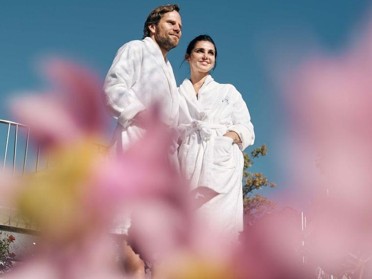 Couple in white bathrobes standing outdoors among flowers under clear blue sky