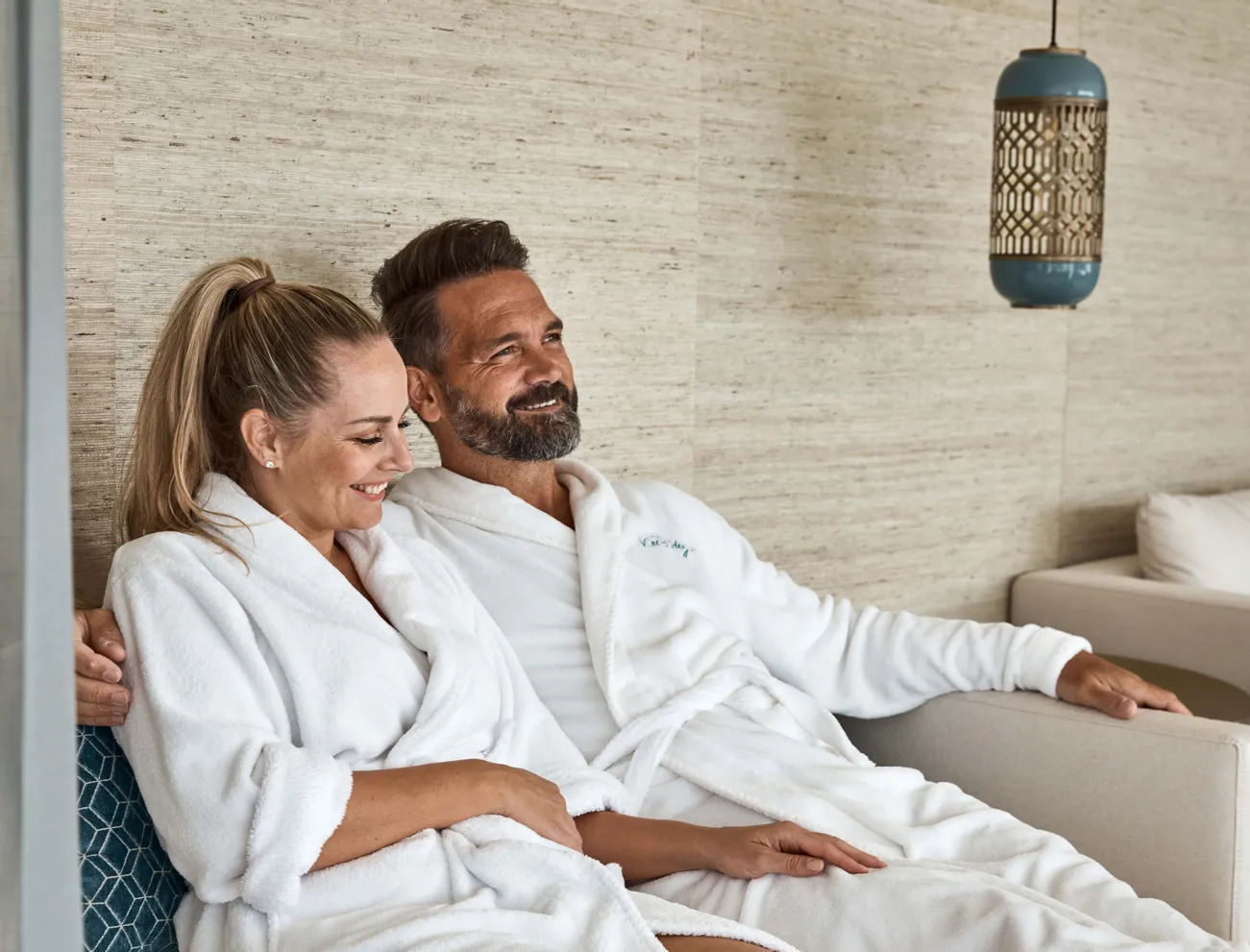 Lächelndes Paar in weißen Bademänteln entspannt auf einem Spa-Loungesofa