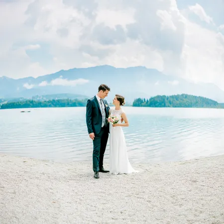 Sposa e sposo in piedi sulla spiaggia del lago con vista montagna sullo sfondo
