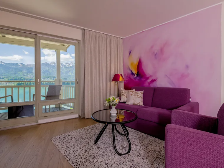 Modernes Zimmer mit violettem Sofa, Blumenkunst, Glastisch und Balkonblick auf Berge und See