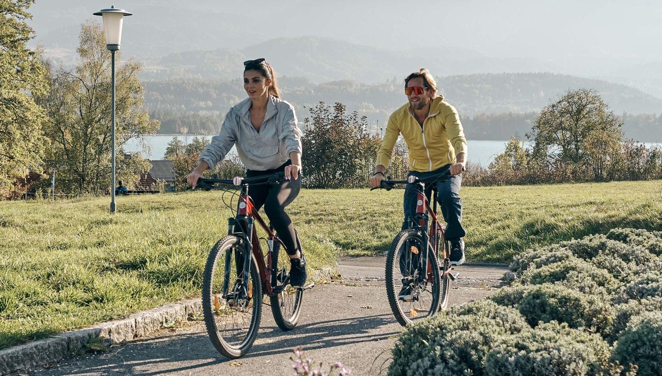 Couple cycling on a scenic path with mountains, lake, and greenery in the background
