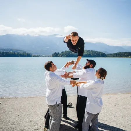 Köche in Uniform werfen Kollegen am See mit Bergkulisse in die Luft
