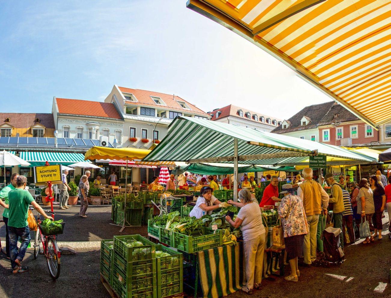 Belebter Bauernmarkt im Freien mit Käufern zwischen Gemüseständen und Blumen unter gestreiften Markisen