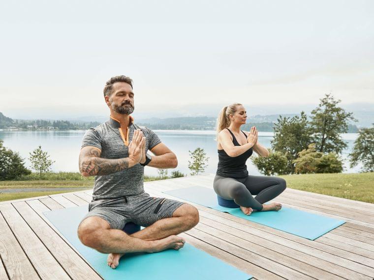 Man and woman practicing seated yoga meditation on mats near a scenic lake