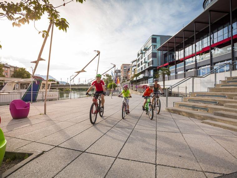 Famiglia in bicicletta sul lungofiume con edifici moderni e barche ormeggiate