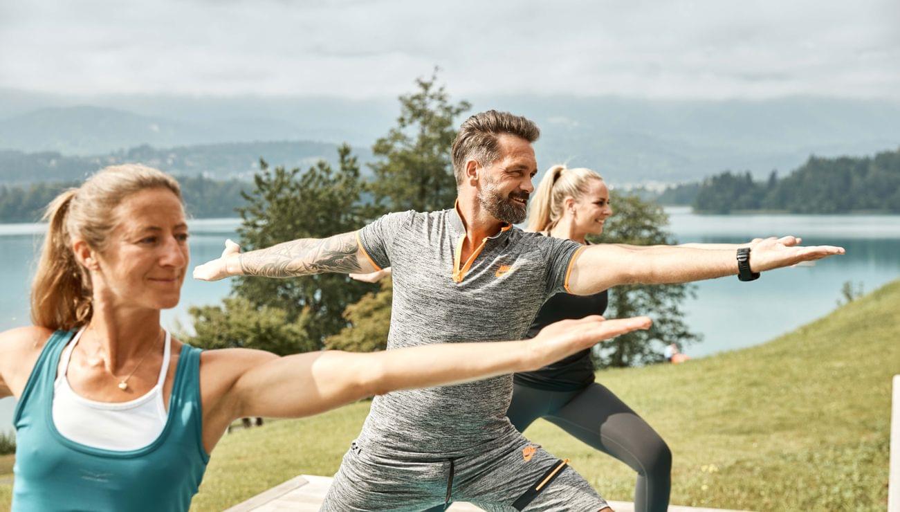 Gruppe beim Yoga im Kriegerpose im Freien am See mit Blick auf Berge