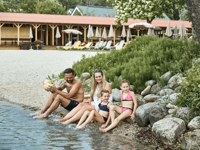 Famiglia felice seduta sulla riva del lago in un resort estivo con cabine sullo sfondo