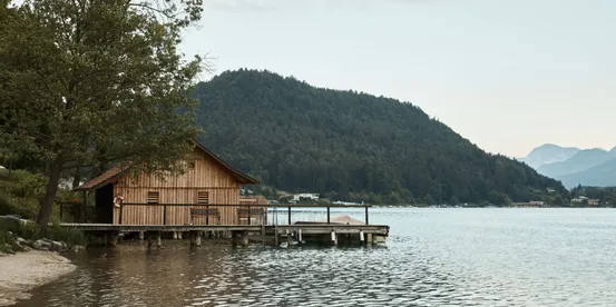 Wooden boathouse on calm lake shore with forested hillside and mountains in background