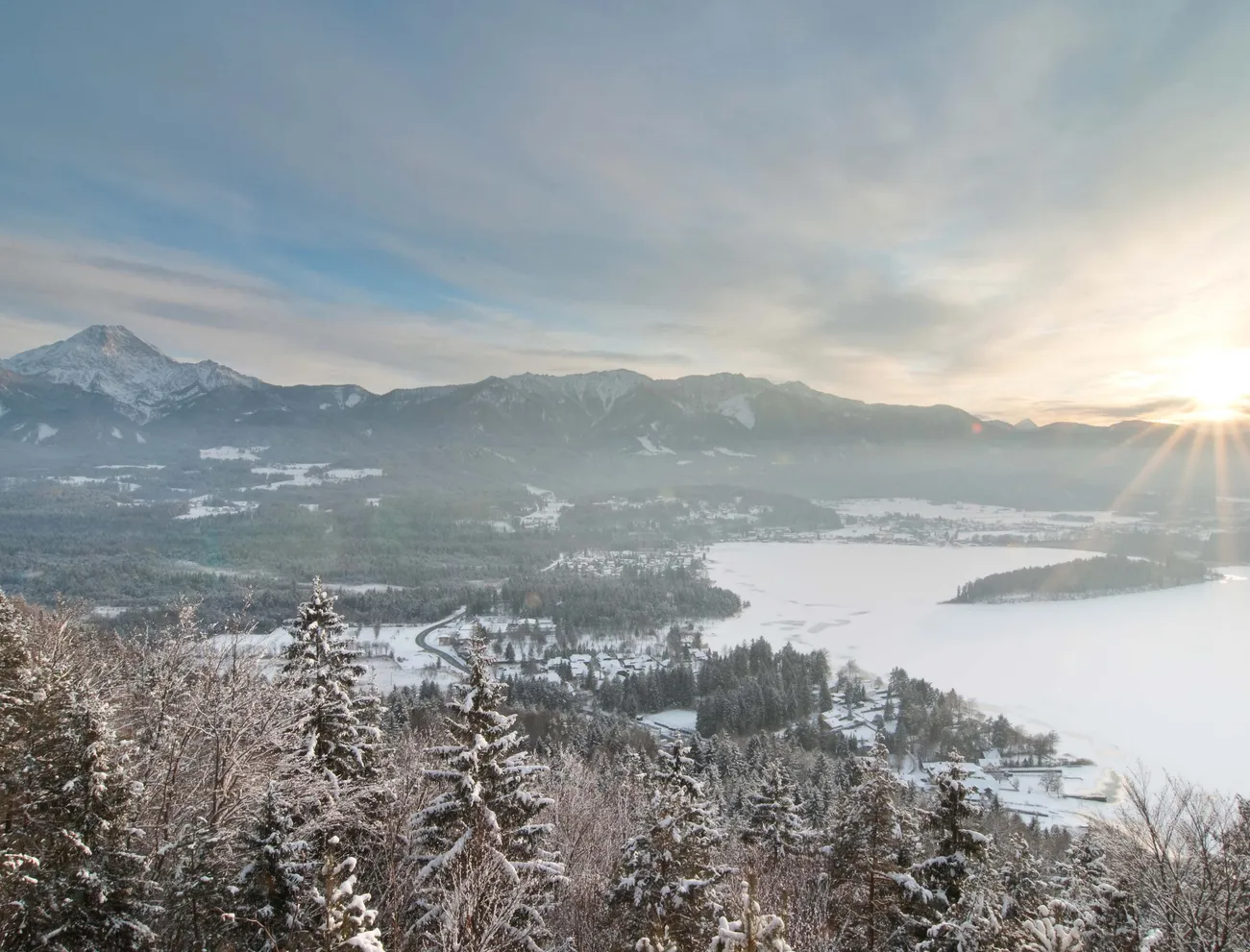Winterlandschaft mit verschneitem Wald, zugefrorenem See und Sonnenaufgang über Bergkette