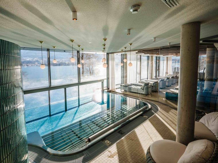 Piscina interna con vista lago, lettini moderni e luci sospese in un resort benessere di lusso