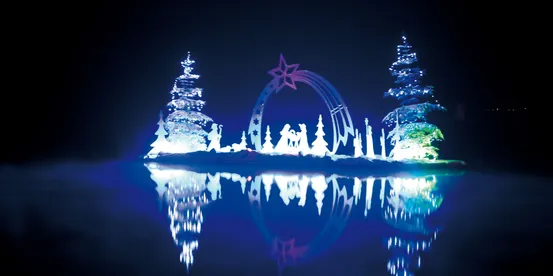 Beleuchtete Weihnachtskrippenszene mit Bäumen und Sternenspiegelung auf dem Wasser bei Nacht