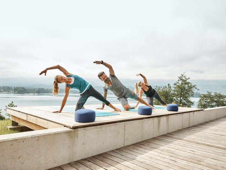 Drei Personen machen Yoga in der Seitstützposition auf einem Holzdeck am See