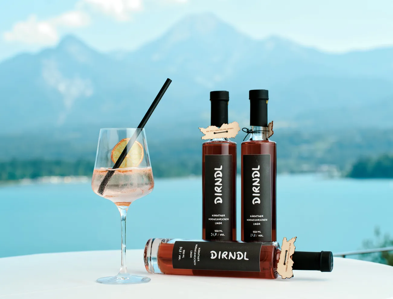Dirndl-Likörflaschen und Cocktailglas mit See und Bergen im Hintergrund