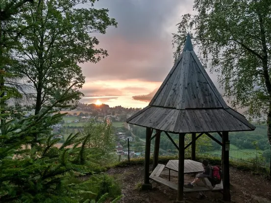 Holzpavillon mit Blick auf ein Tal bei Sonnenuntergang, Wanderer sitzt daneben
