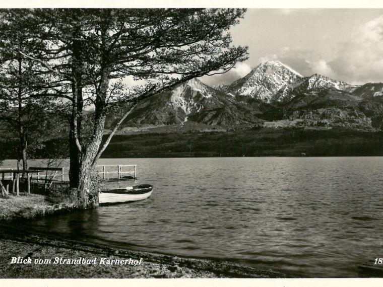 Lake view with dock, boat, trees and snow-capped mountains at Strandbad Karnerhof