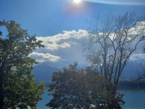 Sonne scheint über einem ruhigen See mit Bäumen und Bergen im Hintergrund