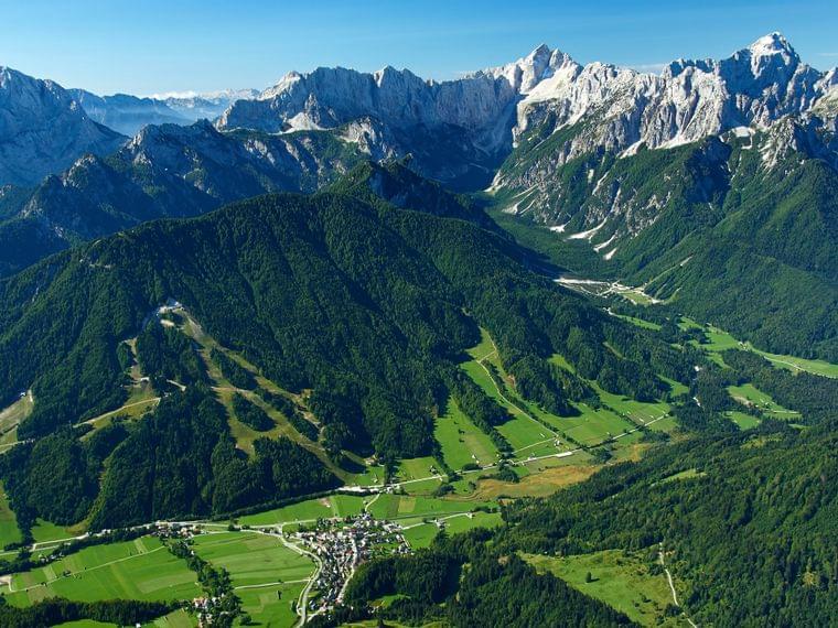Vista aerea di un villaggio alpino in una valle verde circondata da colline boschive e montagne innevate