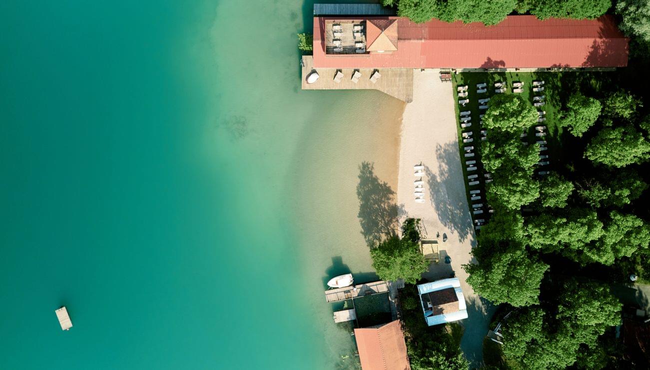 Aerial view of lakeside beach resort with sun loungers, turquoise water and lush green trees