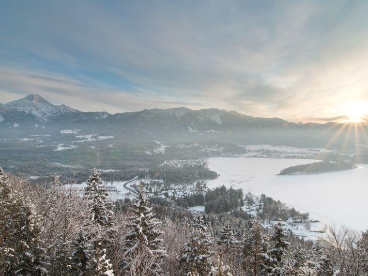 Verschneite Alpenlandschaft mit Wald, zugefrorenem See und aufgehender Sonne über Bergen
