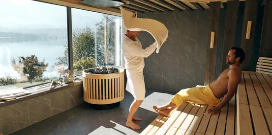 Mann führt Sauna-Aufguss-Ritual mit Handtuch in modernem Spa mit Seeblick durch