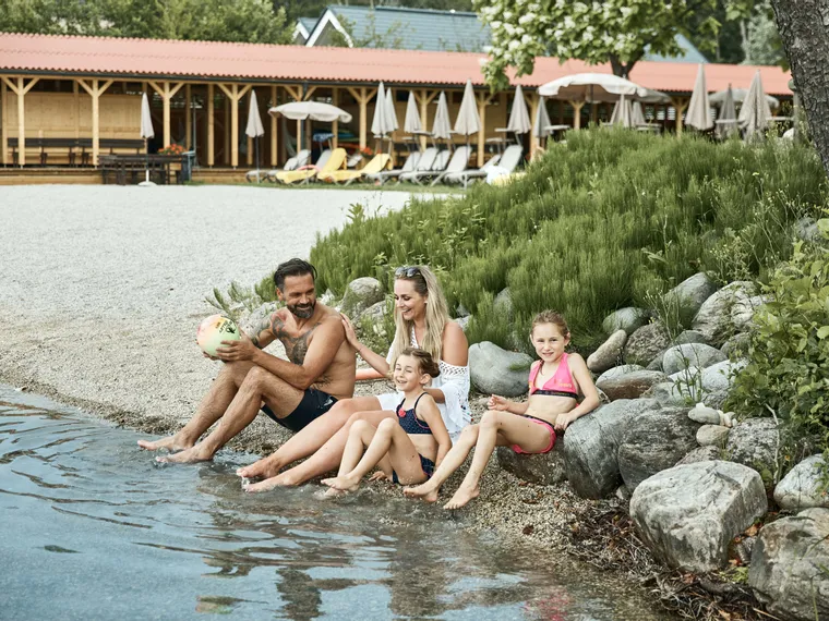 Glückliche Familie sitzt am Seeufer, Kinder planschen mit den Füßen im Wasser im See-Resort