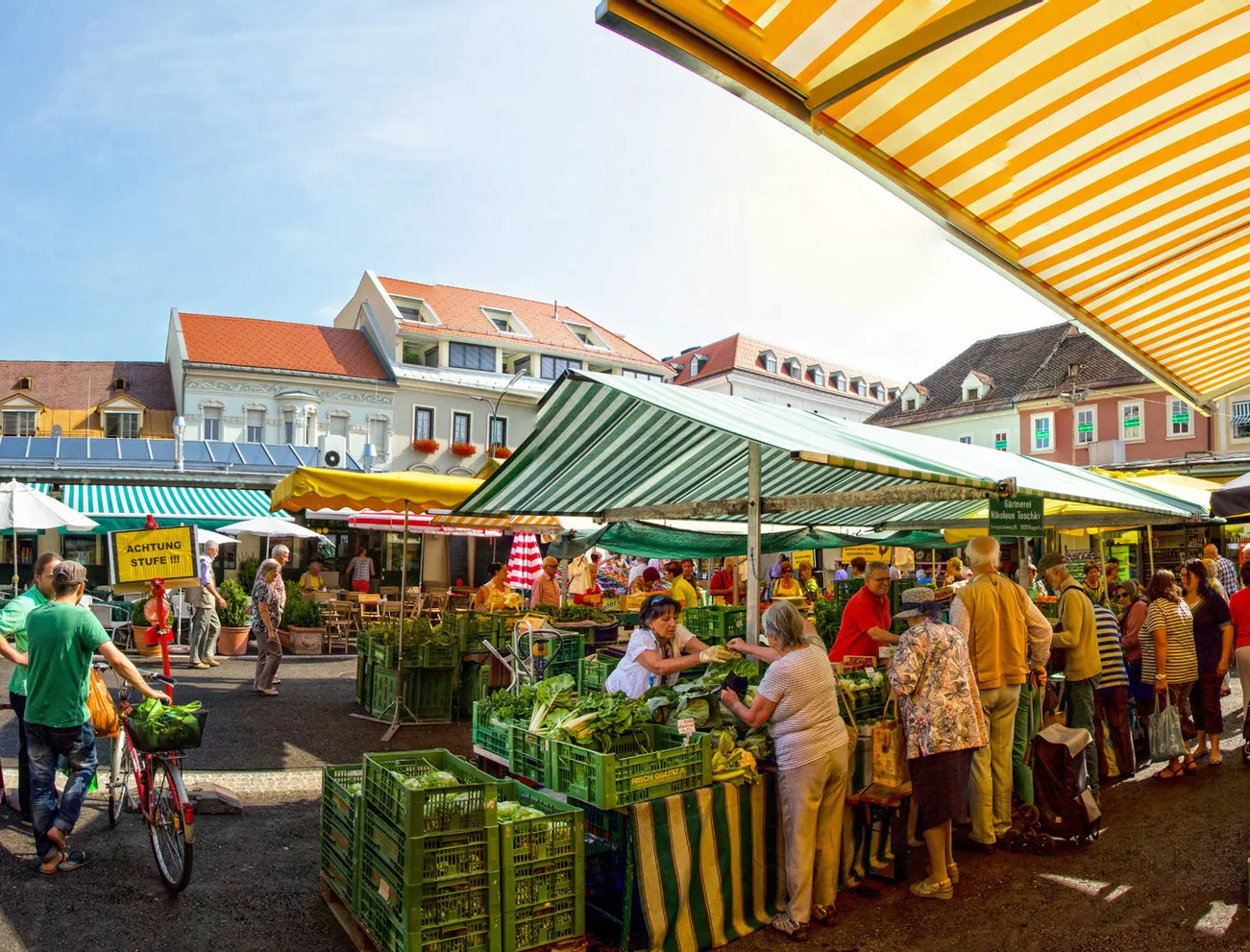 Belebter Bauernmarkt im Freien mit Käufern zwischen Gemüseständen und Blumen unter gestreiften Markisen