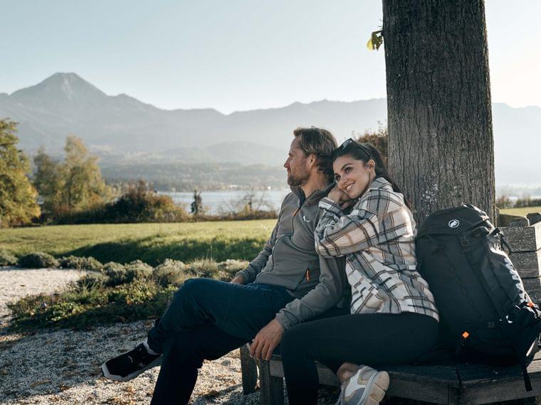 Paar entspannt auf Bank mit Wanderausrüstung unter Baum mit Blick auf Bergsee