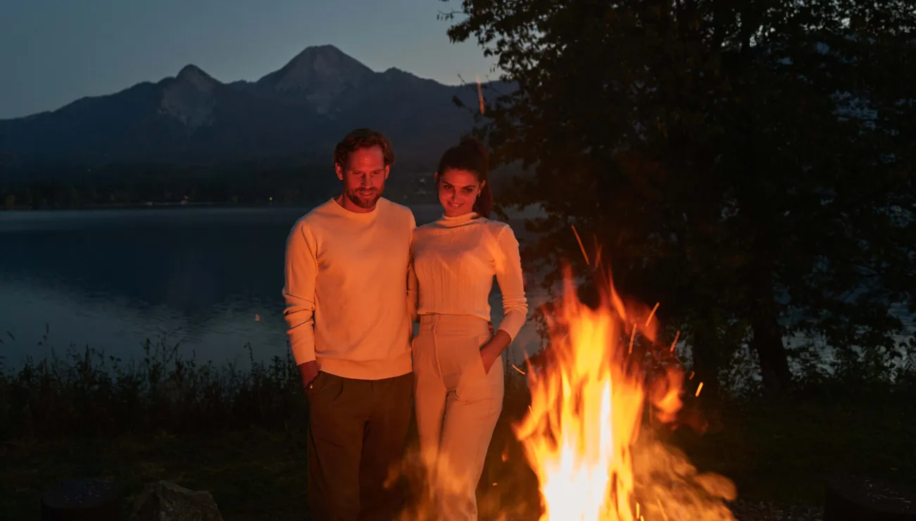 Paar steht bei einem Lagerfeuer an einem See mit Bergblick in der Dämmerung