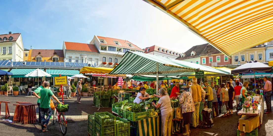 Belebter Bauernmarkt im Freien mit Käufern zwischen Gemüseständen und Blumen unter gestreiften Markisen