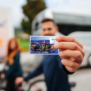 Mann hält Kärnten Erlebnis Card mit Stadtansicht vor einem Reisebus