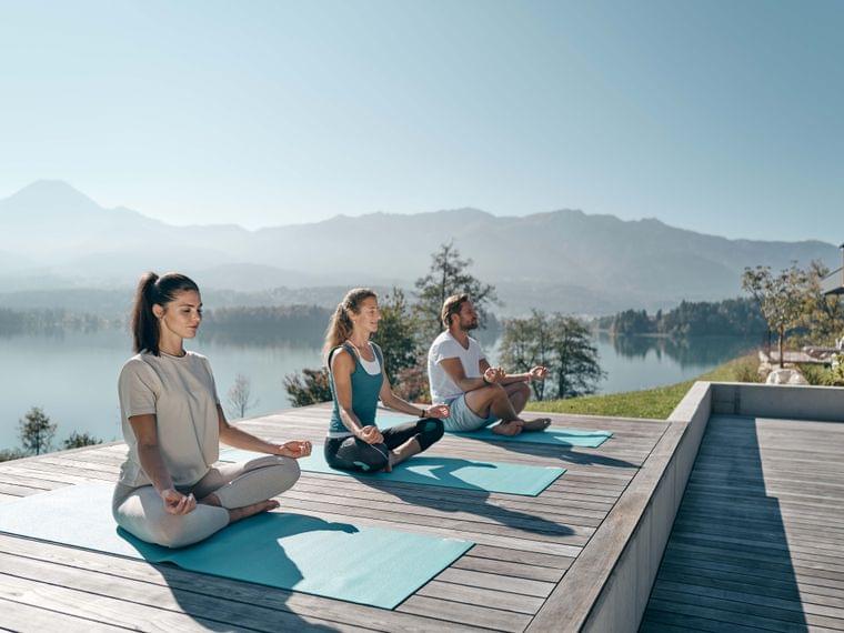 Drei Personen meditieren draußen auf Yogamatten am See mit Bergblick
