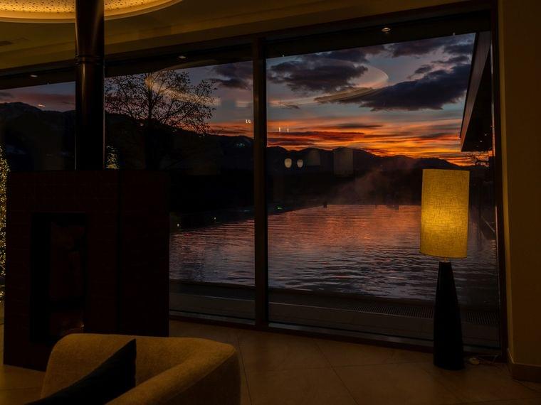 Gemütliche Lounge mit Kamin und Sonnenuntergangsblick über Infinity-Pool durch große Glasfront
