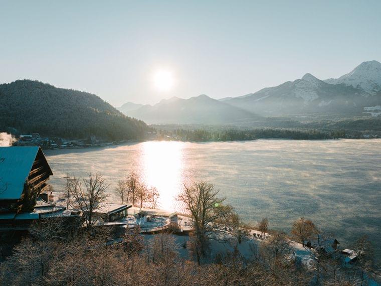 Alba invernale su lago alpino con baita innevata e nebbia sopra l'acqua