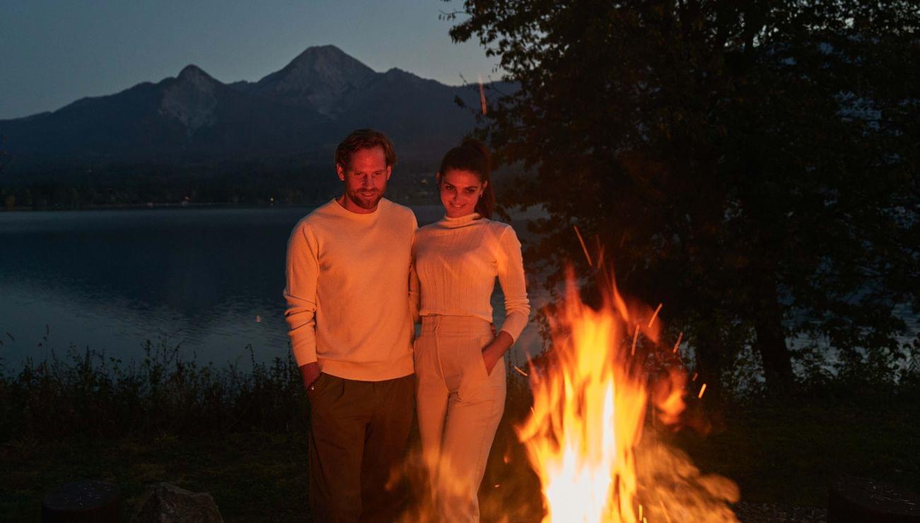 Paar steht bei einem Lagerfeuer an einem See mit Bergblick in der Dämmerung