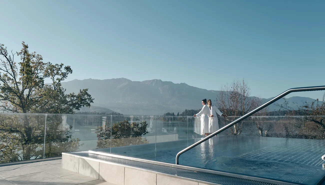 Couple in bathrobes by infinity pool overlooking lake and mountains at luxury wellness resort
