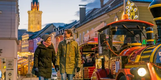 Paar geht an festlichem Straßenbähnchen auf dem Weihnachtsmarkt in abendlicher Stadtszene vorbei