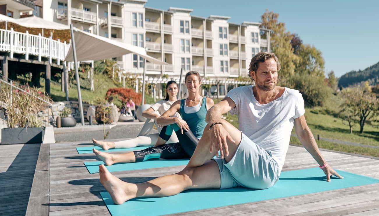 Gruppe macht Yoga im Freien auf Matten vor einem Wellnesshotel mit Bergkulisse