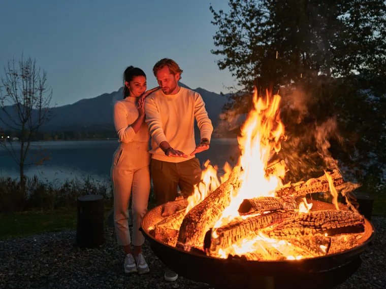 Couple warming hands by a fire pit near a lake at dusk with mountains in the background