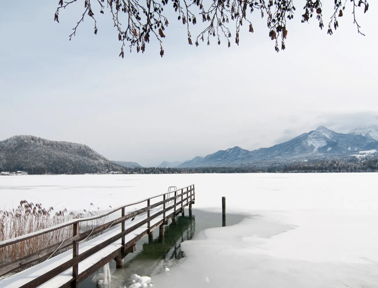 Verschneiter Holzsteg auf zugefrorenem Alpensee mit Blick auf verschneite Berge