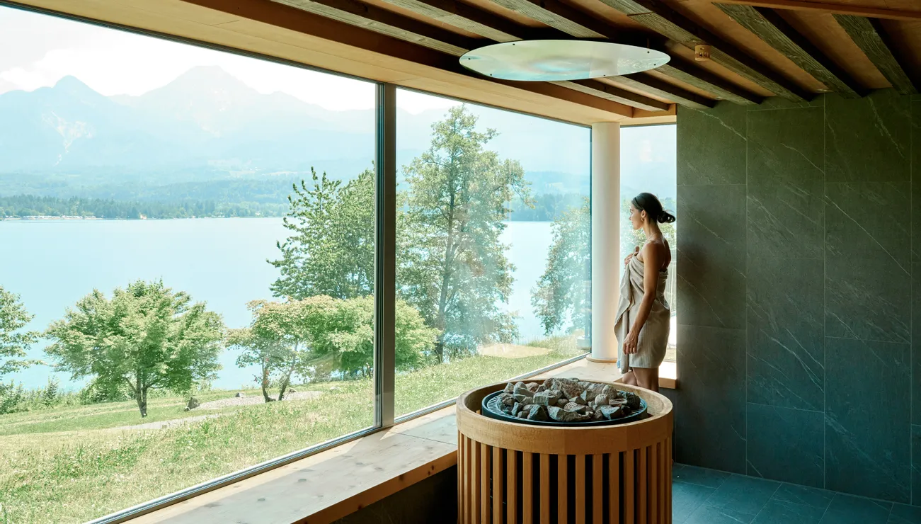 Frau in Saunatuch genießt See- und Bergblick durch großes Fenster in der Sauna