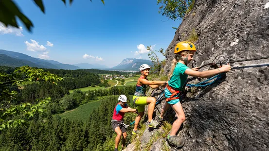 Familie klettert auf Klettersteig bei sonnigem Wetter mit Wald und Tal im Hintergrund
