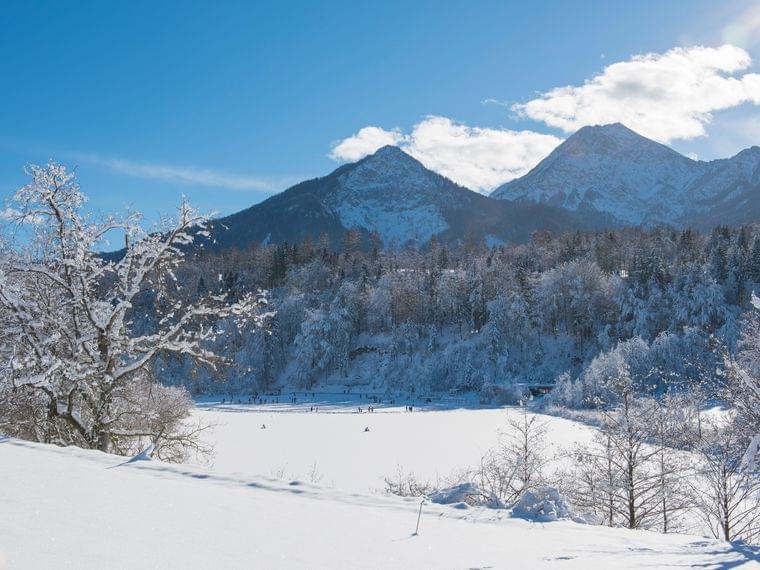 Verschneiter Wald und zugefrorener See mit Menschen vor Alpenbergen unter klarem blauen Himmel