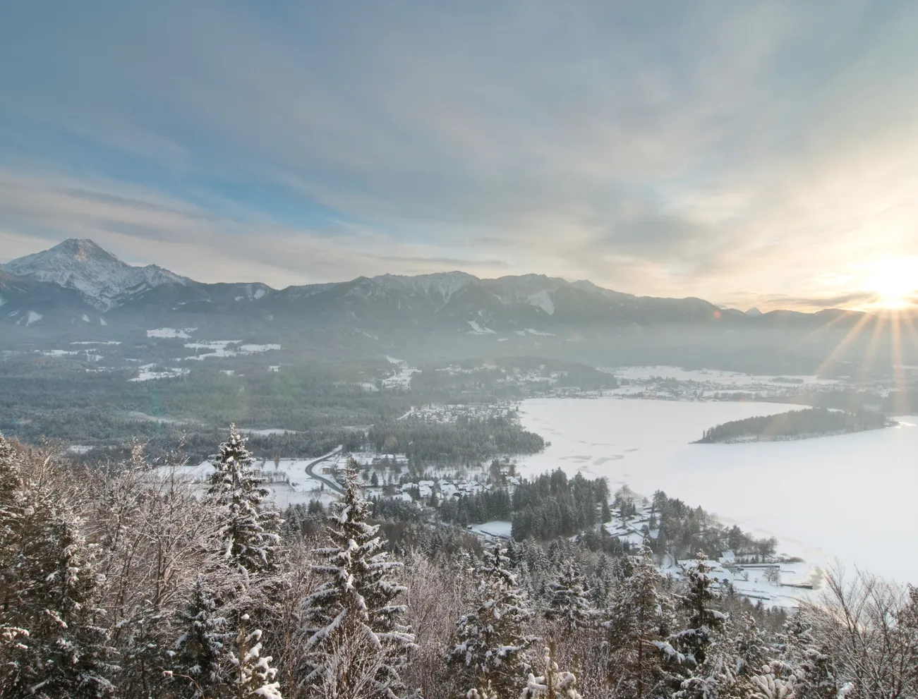 Verschneite Alpenlandschaft mit Wald, zugefrorenem See und aufgehender Sonne über Bergen