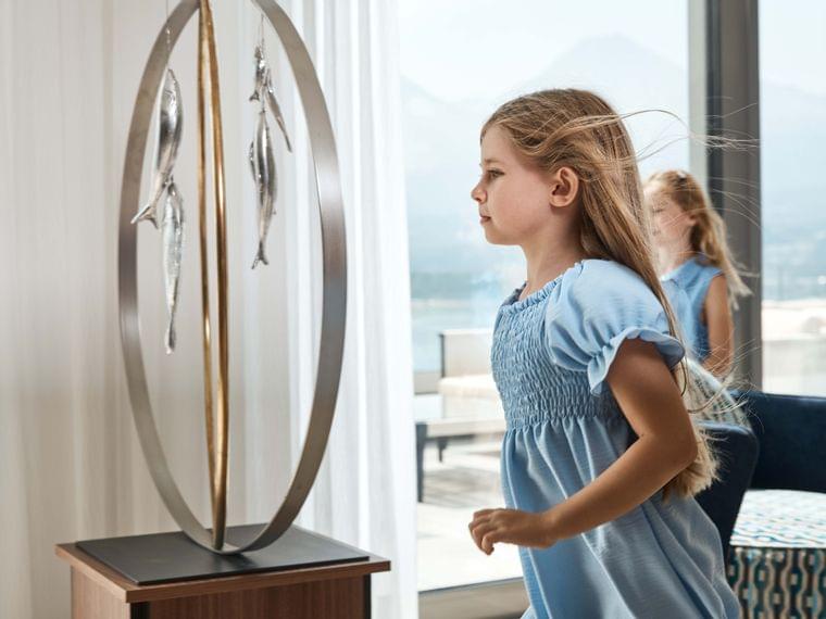 Young girl in blue dress looking at modern metal fish sculpture indoors