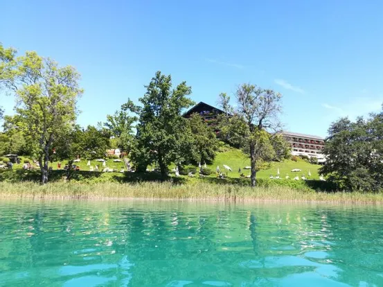Ferienresort am See mit grünem Hügel, Bäumen und Liegestühlen unter blauem Himmel