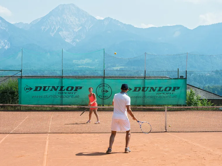 Two people playing tennis on a clay court with mountains in the background