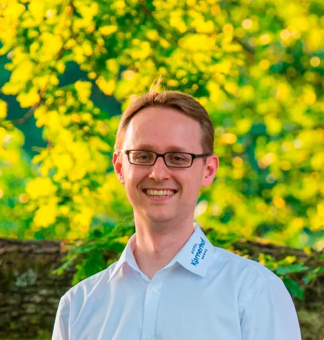 Uomo sorridente con camicia bianca e logo Hotel Karwendel davanti a fogliame verde
