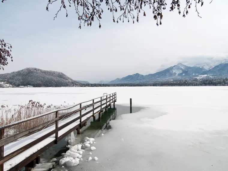 Verschneiter Holzsteg auf zugefrorenem Alpensee mit Blick auf verschneite Berge