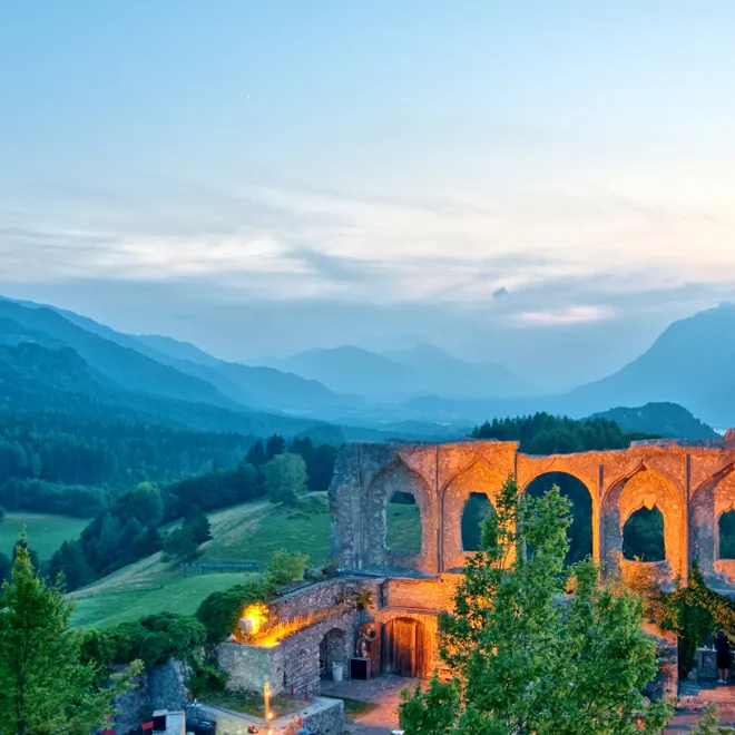 Beleuchtete Burgruine bei Sonnenuntergang mit Alpen im Hintergrund